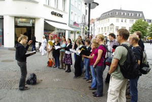 Chor in Straßenkleidung singt in Fußgängerzone, Chorleiterin Ines Kaun dirigiert