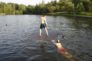 Mann stehend auf Holzplattform im See, zwei Männer im Wasser schieben die Plattform