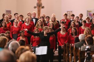 Chor in mehreren Reihen, davor Chorleiterin Ines Kaun zum Publikum gewendet, die Arme ausbreitend