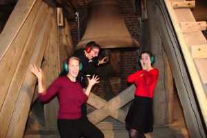 Drei Personen mit Lärmschutzkopfhörern schneiden Grimassen vor einer Kirchenglocke