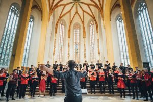 Blick von unten auf singenden Chor während eines Konzertes, Chorlieter Fabian Pasewald im Vordergrund und das Gewölbe über dem Altarraum im Hintergrund
